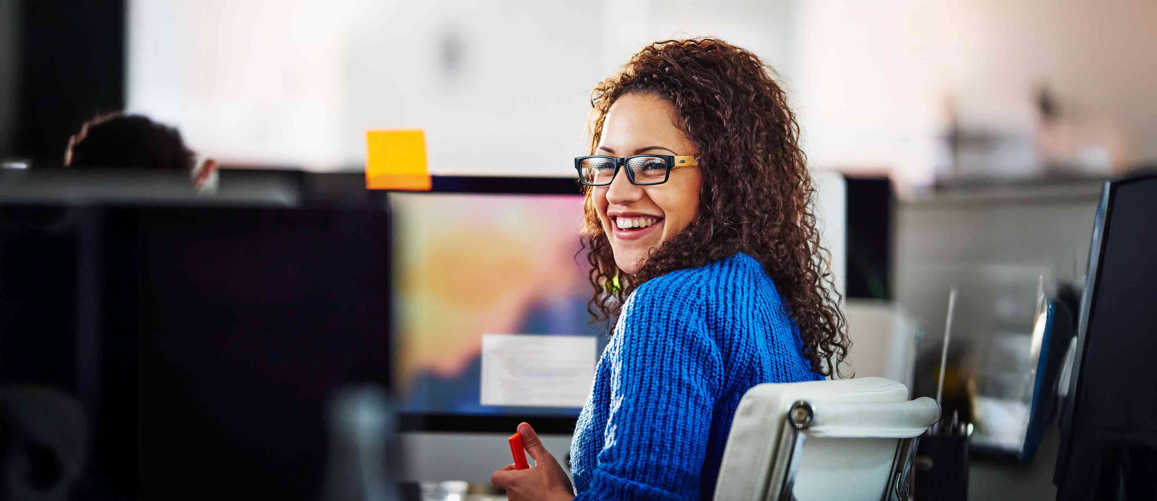 Woman sits at computer desk conversing with a colleague